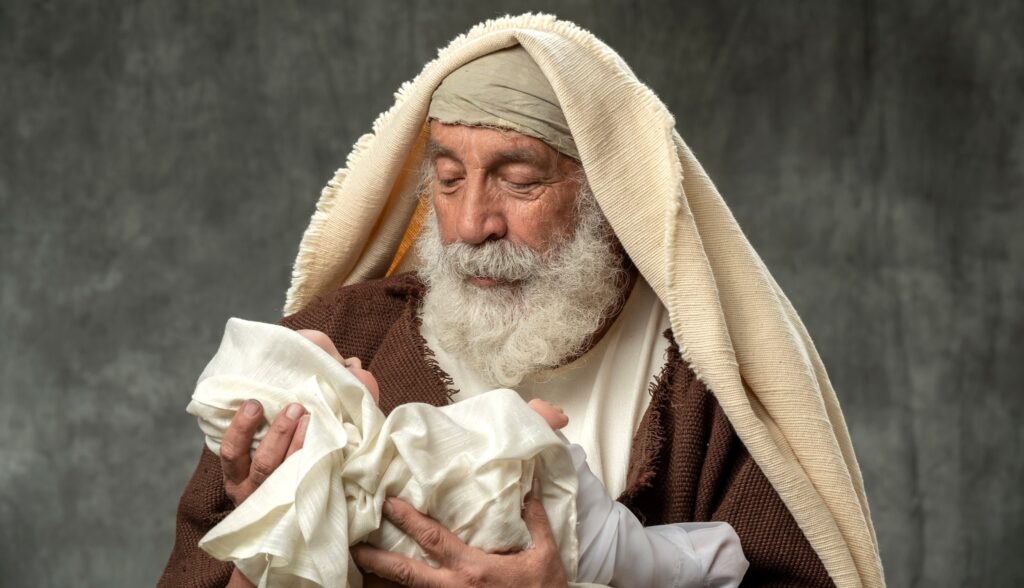 Simeon holding baby Jesus against a dar gray backgroung